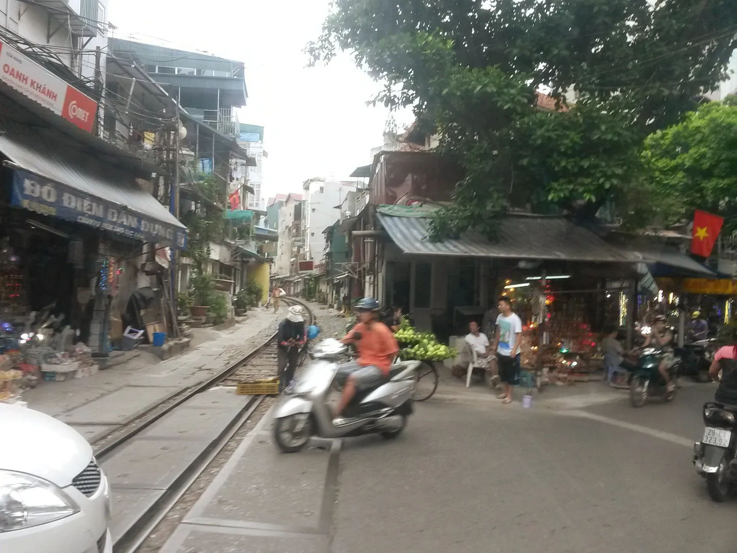 Ulica w Starej Dzielnicy Hanoi z torami kolejowymi biegnącymi przez środek, motocyklistami i lokalnymi straganami po bokach.