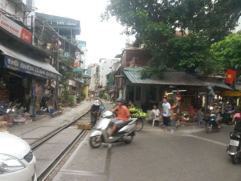Ulica w Starej Dzielnicy Hanoi z torami kolejowymi biegnącymi przez środek, motocyklistami i lokalnymi straganami po bokach.
