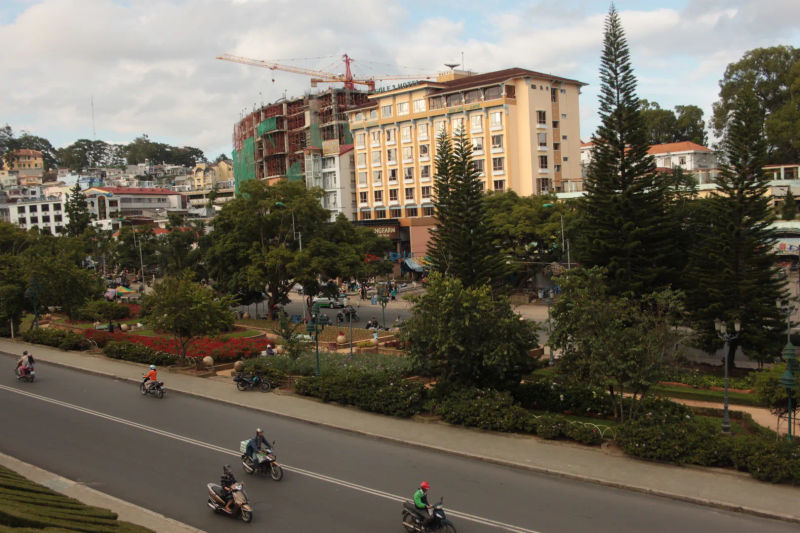 Widok na centralny plac w mieście Da Lat, Wietnam, z zielonymi terenami, wysokimi budynkami i motocyklistami na drodze.