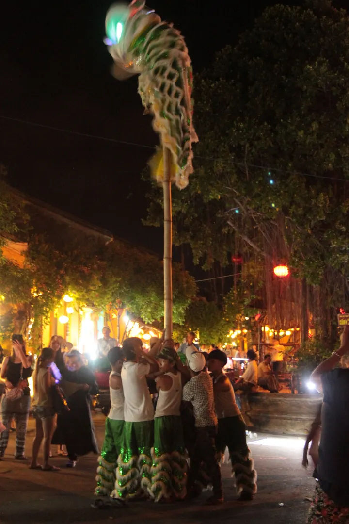 Wieczorna scena w Hoi An z kolorowymi lampionami i tłumem spacerujących ludzi, otoczona bujną roślinnością.