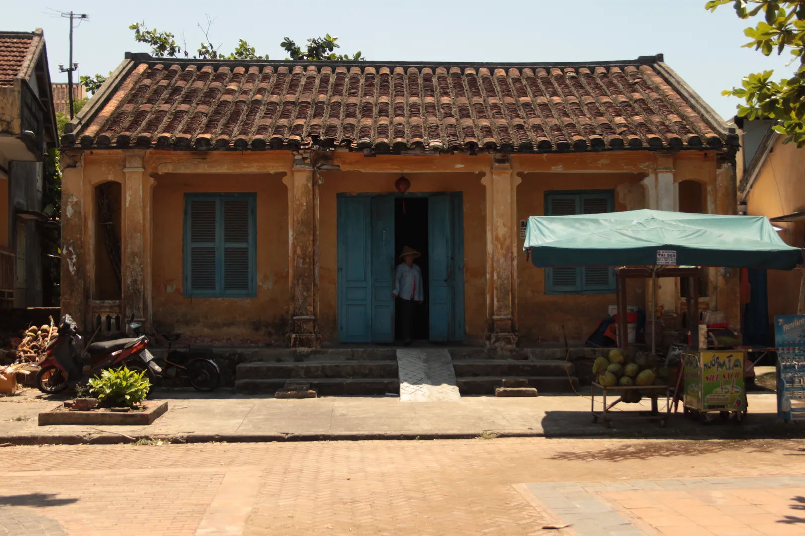 Niewielki, tradycyjny dom z żółtą fasadą i niebieskimi okiennicami, przed którym stoi kram uliczny, Hoi An.