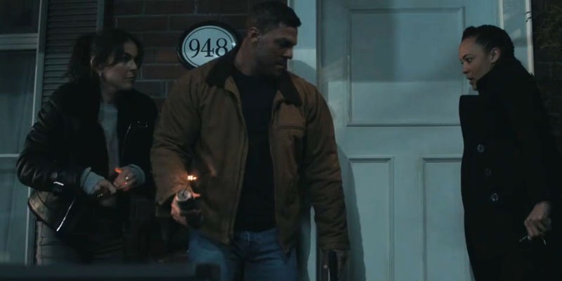 Serinda Swan as Dixon and Maria Sten as Neagley watching Alan Ritchson as Reacher as he holds a lit pipe bomb outside the door of a suburban house