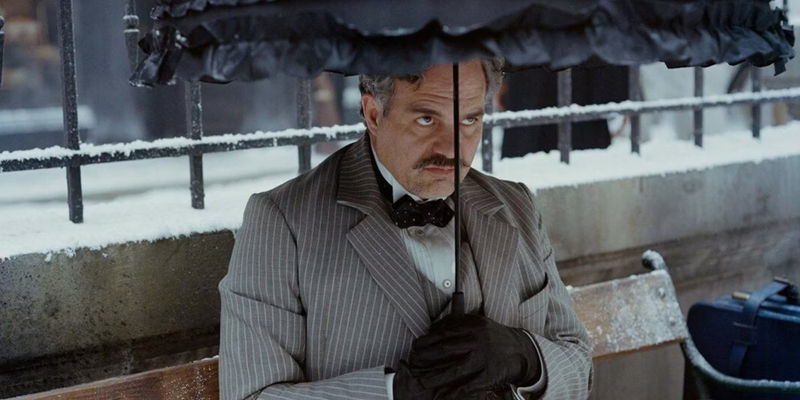 Mark Ruffalo as Duncan Wedderburn, wearing a grey pinstripe suit and sitting under an umbrella, in Poor Things