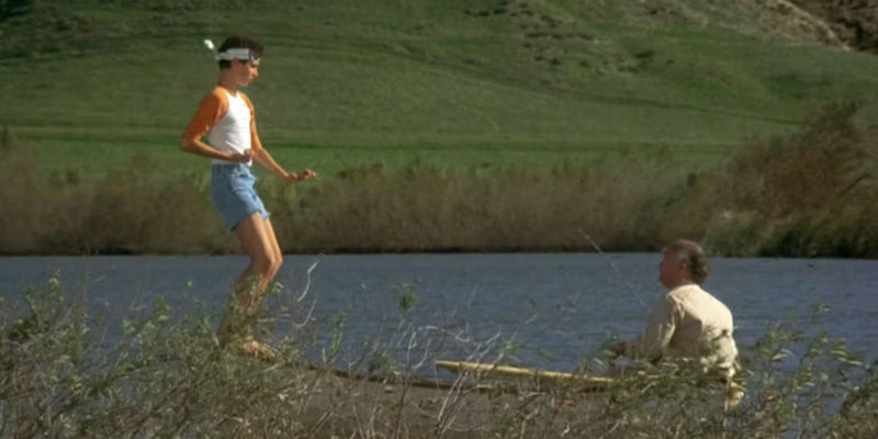Daniel balances on a boat while Miyagi sits and watches in The Karate Kid