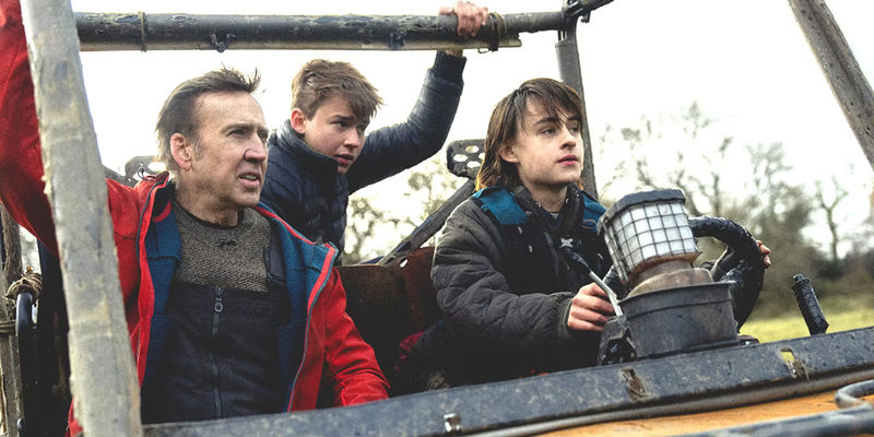 Nicolas Cage and two kids in winter attire riding in an open-air vehicle through a forest setting in Arcadian