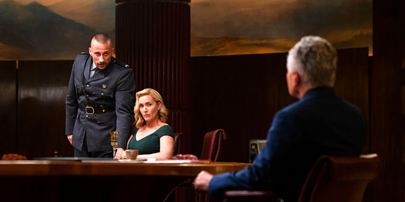 Matthias Schoenaerts and Kate Winslet in a government briefing in The Regime