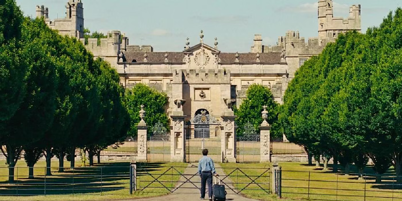 Oliver stands at the gates of Saltburn with his suitcase in Saltburn.