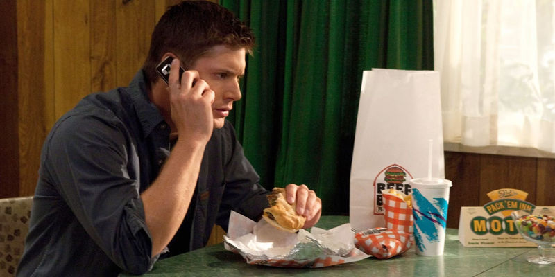 Jensen Ackles as Dean Winchester sitting at a table and eating a burger while talking on the phone in Supernatural