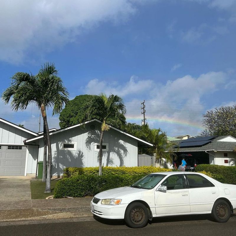 A white sedan is parked in front of a house with a garage under a partly cloudy sky. There are palm trees, and a faint rainbow is visible in the background. No people are discernible in the image.