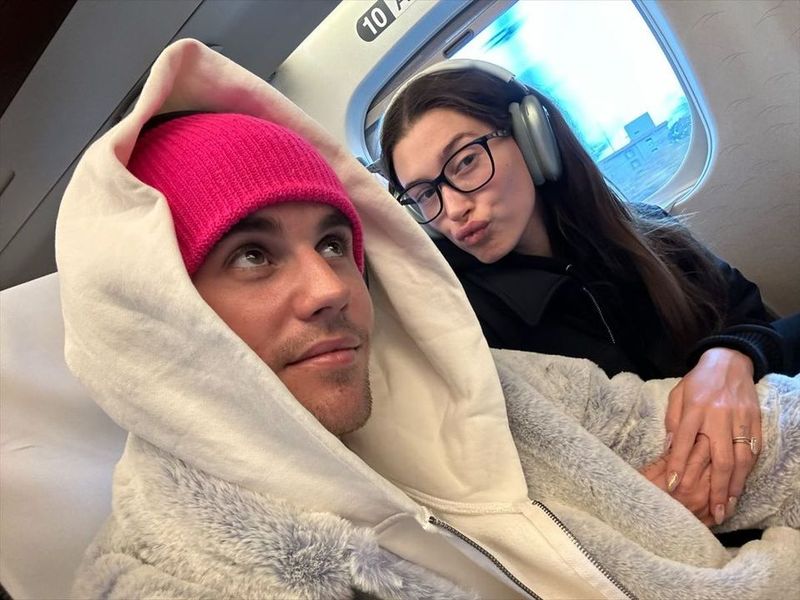 Justin Bieber in a pink beanie beside a woman with headphones on a train, making a playful face.