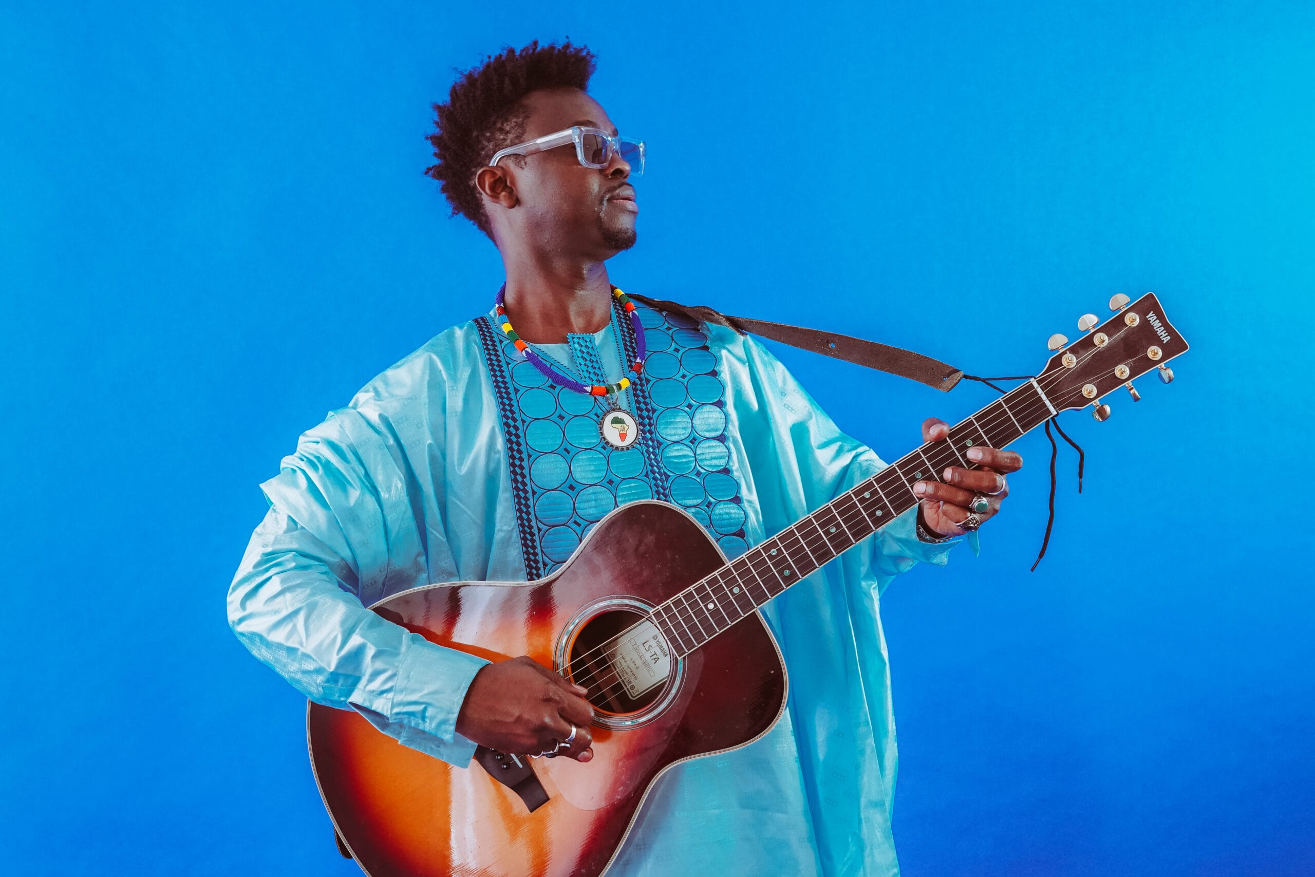 Homme en tenue traditionnelle africaine avec sa guitare sur fond bleu.