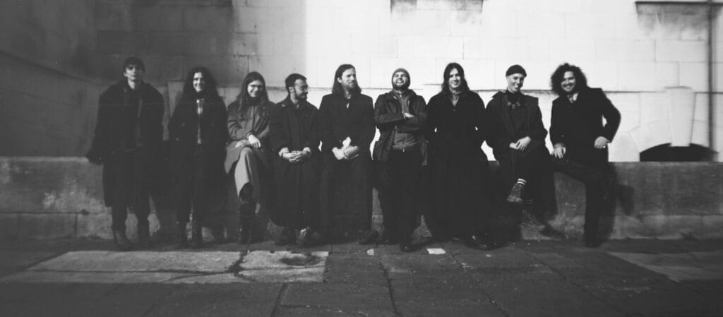 neuf musiciens et musiciennes en noir et blanc assis sur un muret.