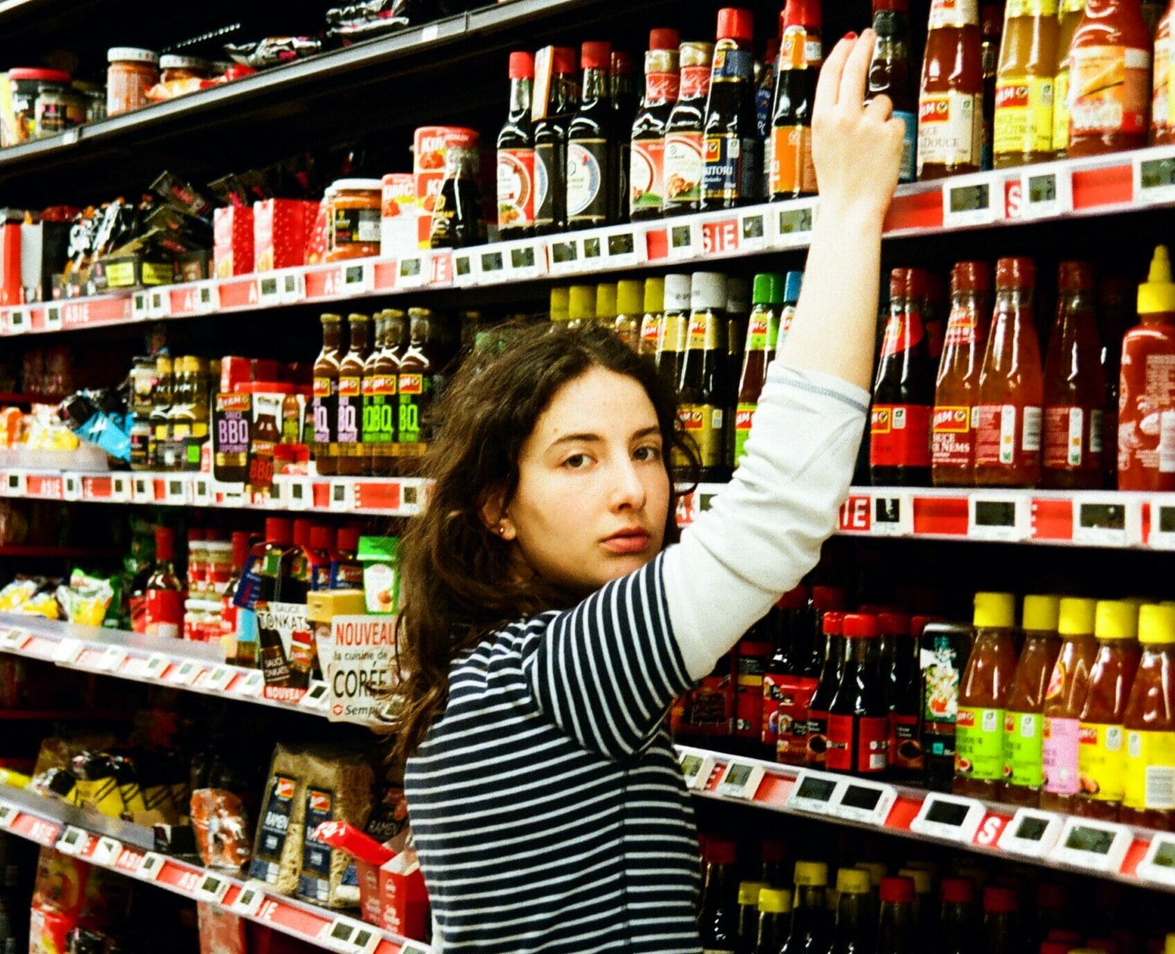 Femme vingtenaire qui cherche à attraper une bouteille de ketchup dans un rayon de supermarché.