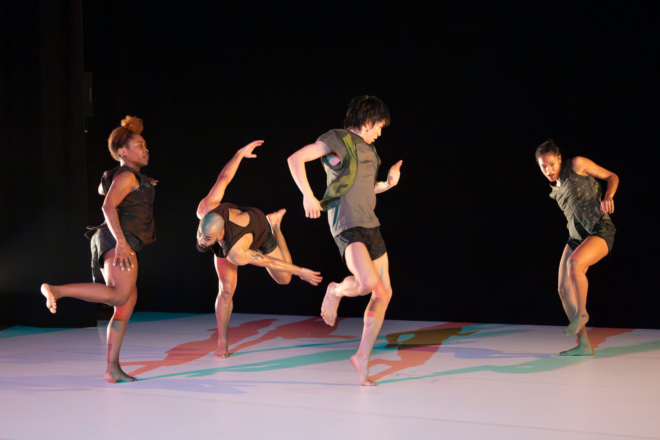 quatre danseurs en short et débardeur ont le pied levé sur une scène.