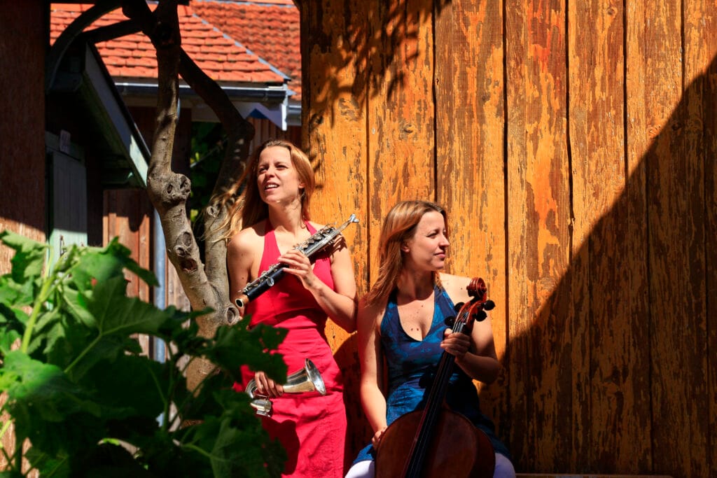 Une clarinettiste et une violoncelliste posent avec leurs instruments dans un jardin au soleil.