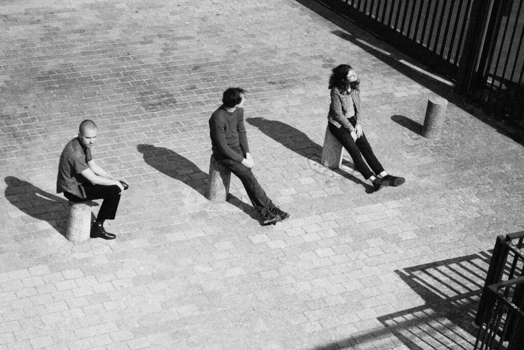 Trois jeunes personnages en noir et blanc posent assis sur des plots urbains face au soleil.