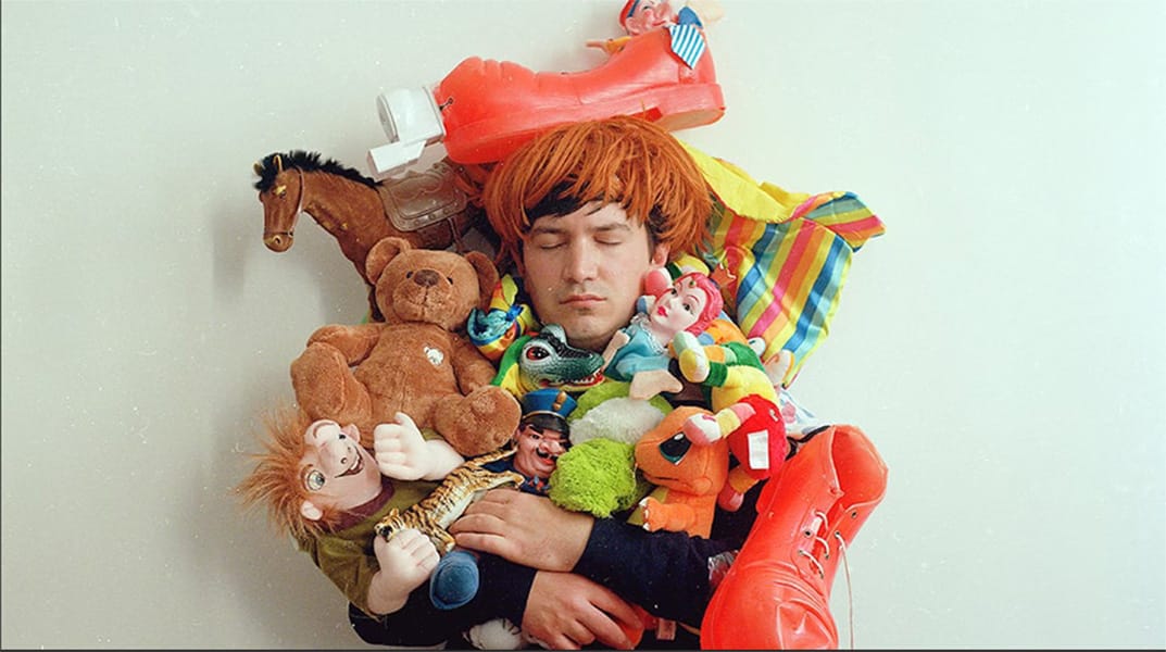 homme avec une perruque rousse entouré d'une multitude de jouets tout autour de sa tête et dans ses bras.