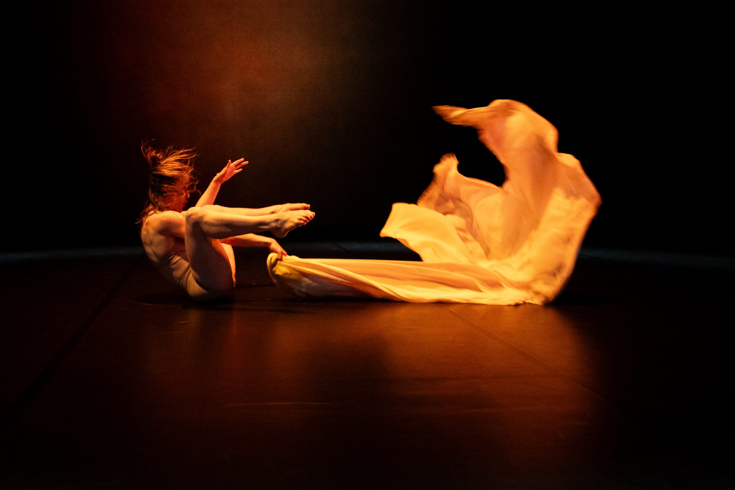 danseuse assise au sol, les jambes relevées secouant un long drap dans une lumière orangée.