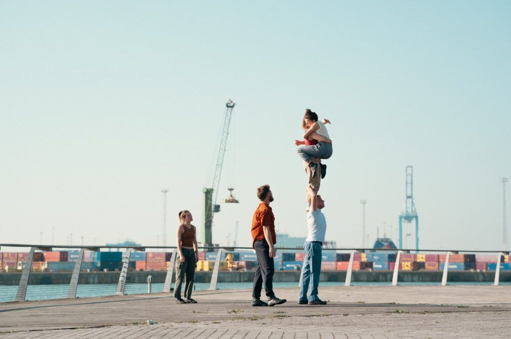 Dans un paysage portuaire, deux acrobates sont grimpées sur les épaules d'un autre.