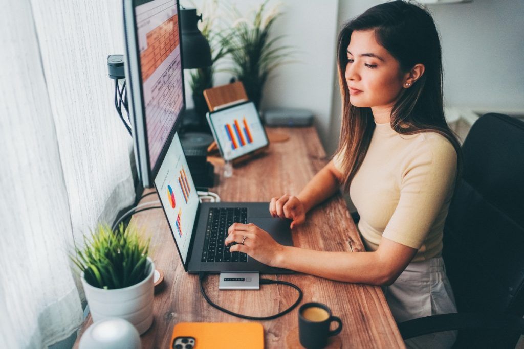 Woman working at a desk with a laptop, external monitor, and tablet displaying colorful charts, with a coffee cup and potted plant nearby.