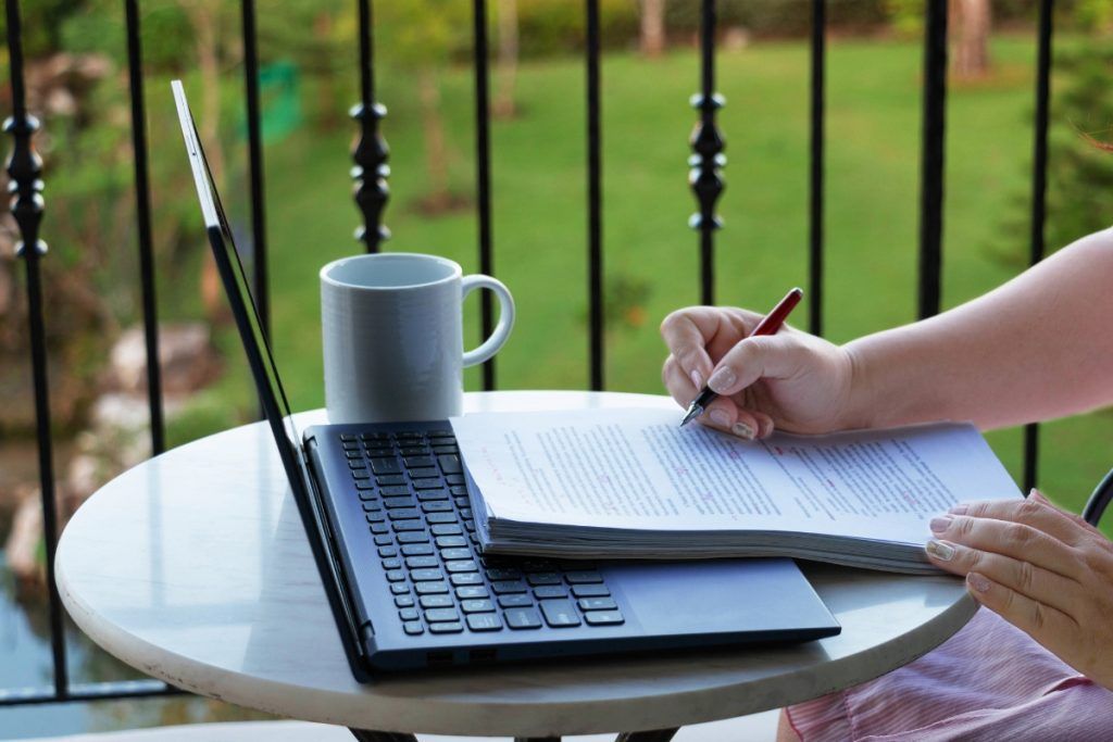 Person working at an outdoor table with a laptop, mug, and open book, writing notes with a pen.