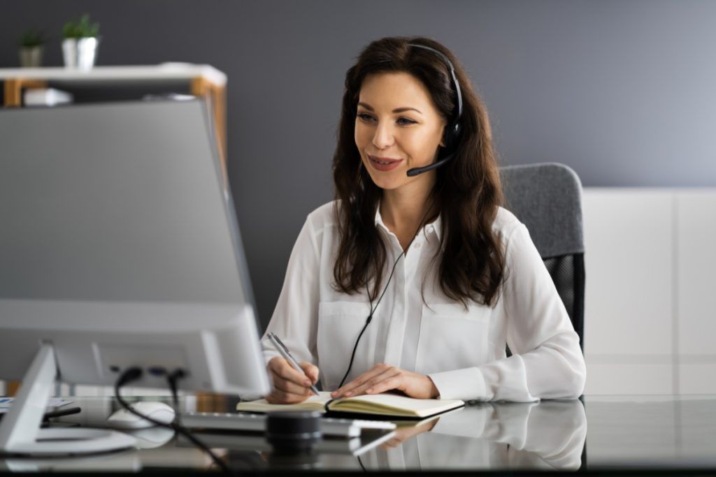 Smiling woman wearing a headset, taking notes in a notebook while participating in a video call on a desktop computer.