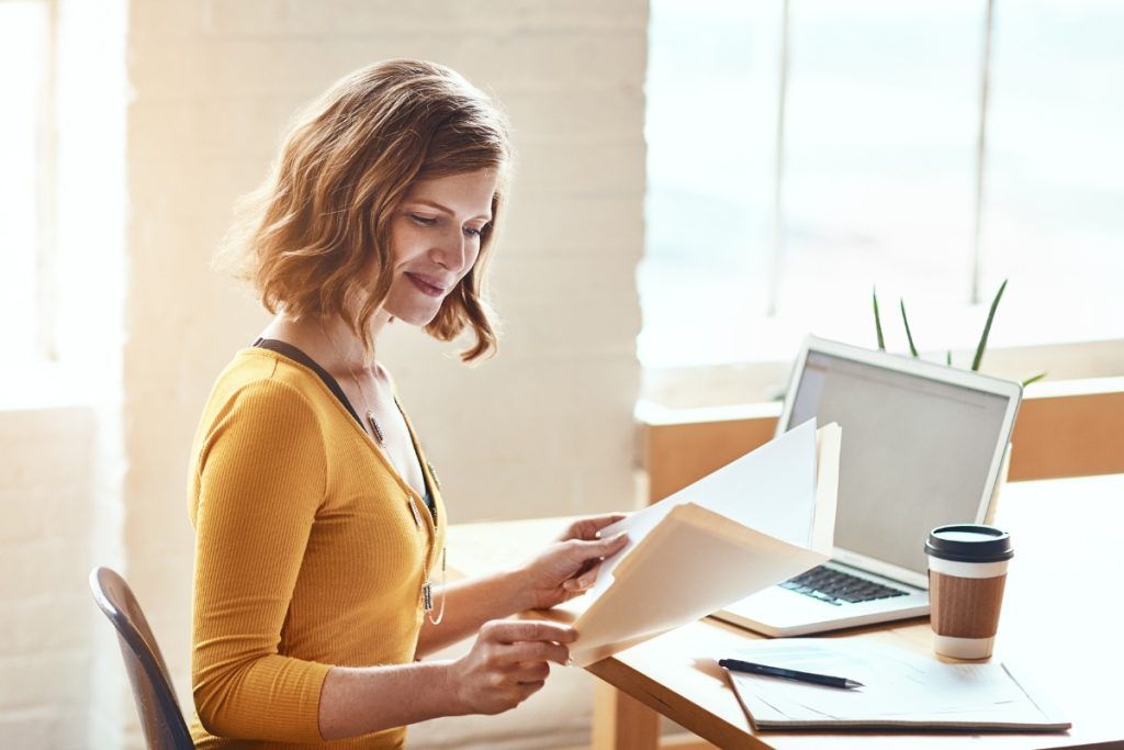 Smiling woman in a yellow top reviewing documents at a desk with a laptop and takeaway coffee cup.