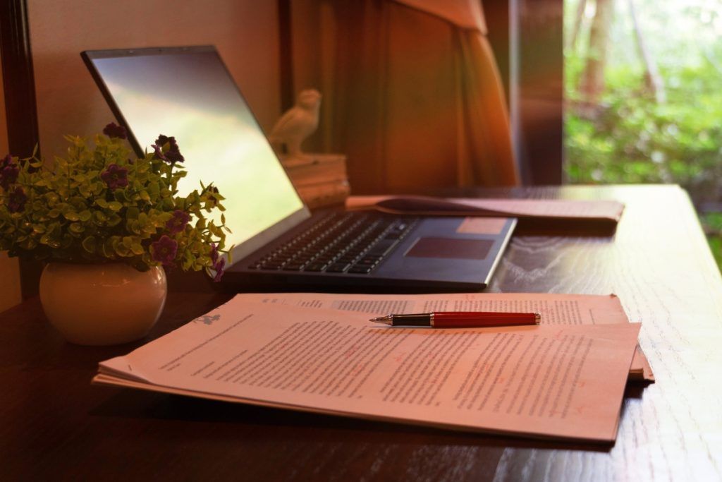 Close-up of a desk with a laptop, potted plant, and printed documents marked with red edits, with a pen resting on top.