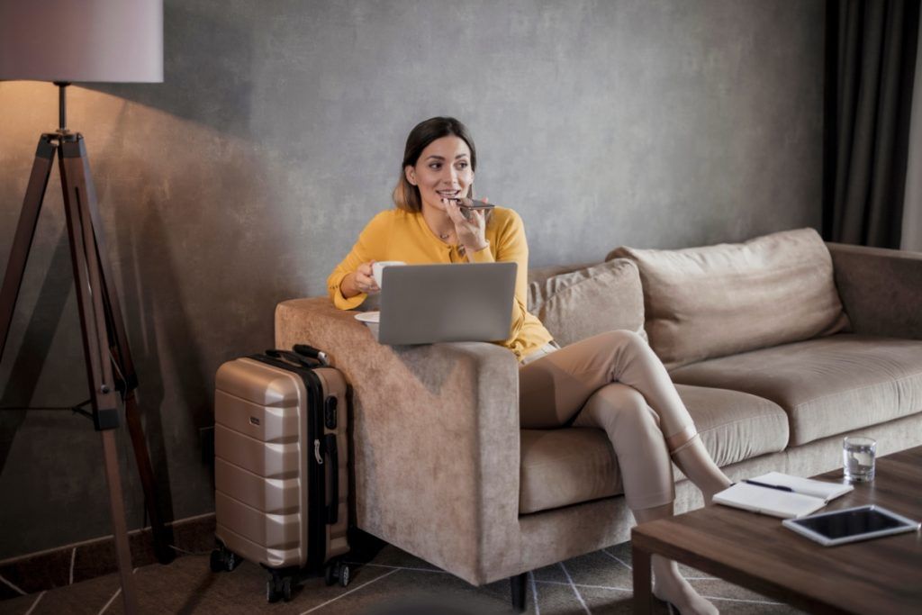 Woman sitting on a couch with a laptop and coffee, speaking into her phone, with a suitcase and notepad on a table nearby.