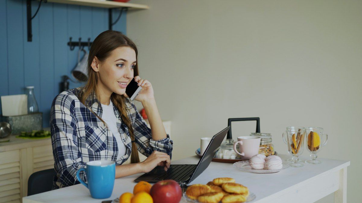 Woman working on a laptop at her kitchen table while on the phone