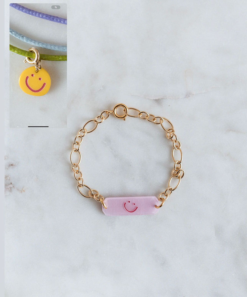 The WALD Berlin Happy ID Yellow bracelet exclusive features a yellow smiley face charm, shown alongside a gold chain bracelet with a pink rectangular charm and red smiley face.