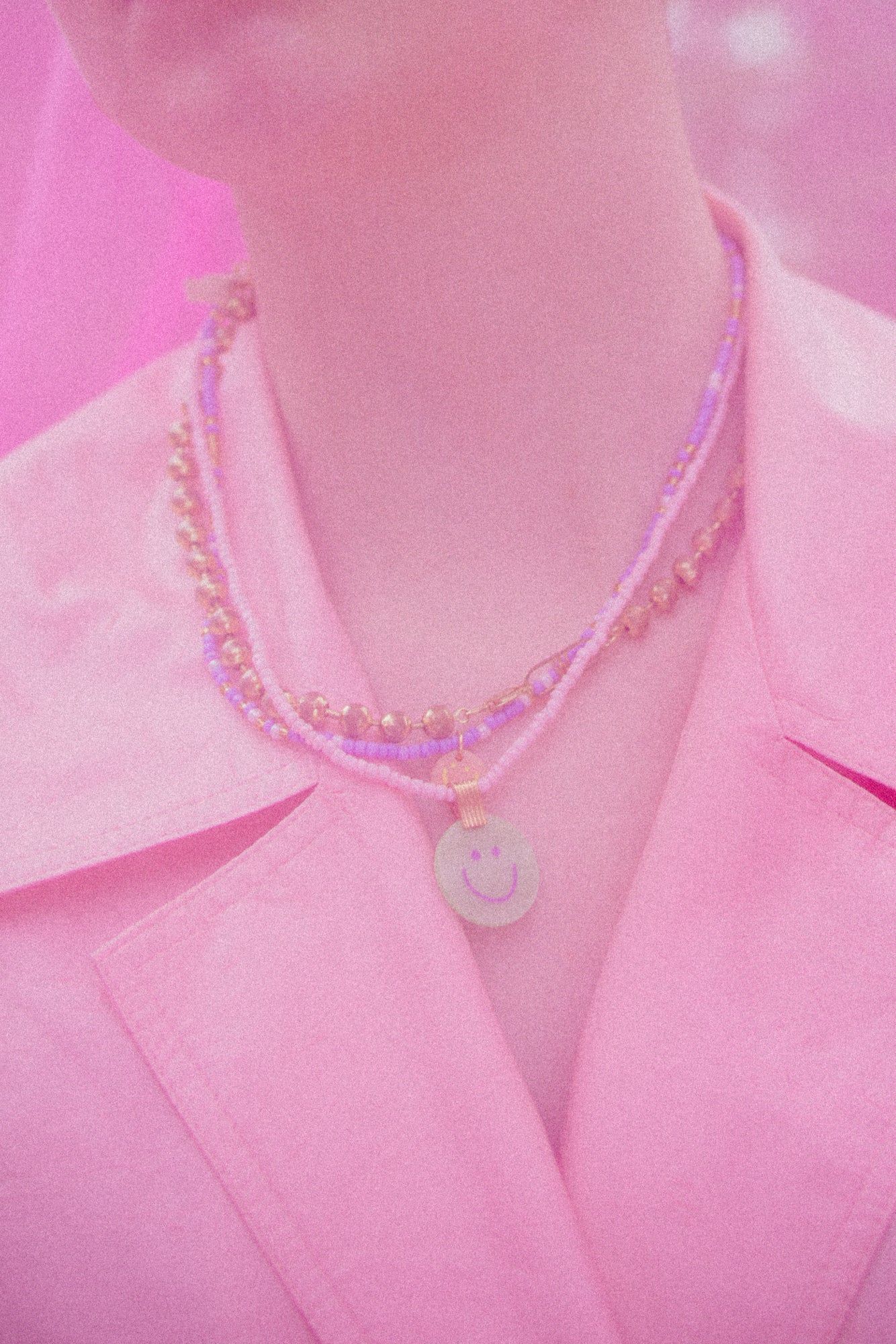 A person wears a pink blazer and layered classic necklaces, including the WALD Berlin Pop Smilie Medallion Peach, shown from the shoulders up.