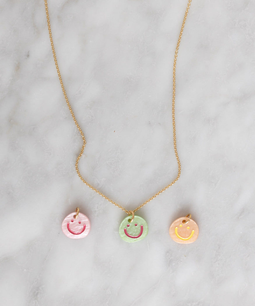 A WALD Berlin Smilie Dude Rose Medaillon Necklace with three hand-painted round pendants featuring smiley faces in pink, green, and yellow, placed on a light marble surface. Crafted from recycled gold and silver, this unique piece is Made in Germany.