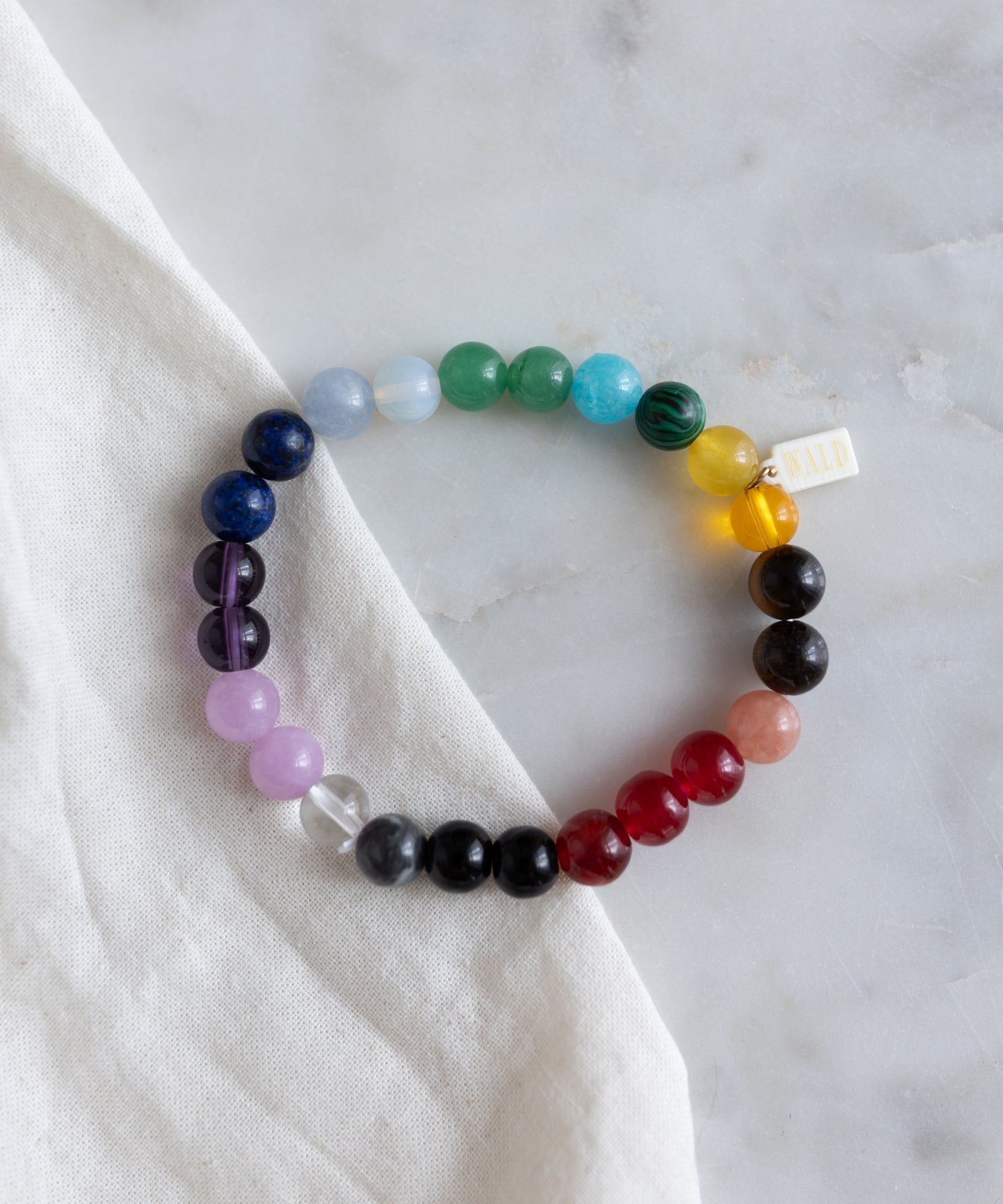 The Over The Rainbow Gemstone Bracelet by WALD Berlin, a multicolored beaded bracelet featuring genuine gemstones arranged in a beautiful gradient of rainbow colors, lies on a white fabric atop a marble surface.