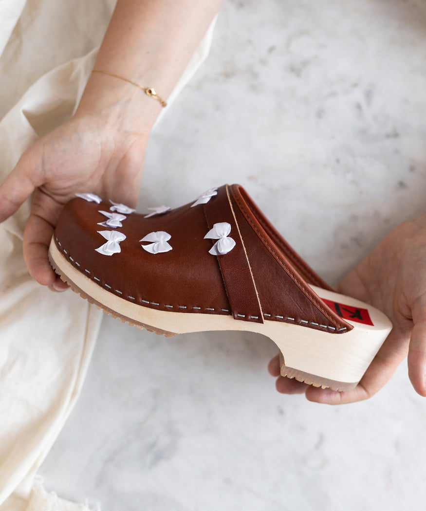 A person holds a brown leather Isla Bonita Clogs by WALD Berlin with white bow accents and a light wooden sole.