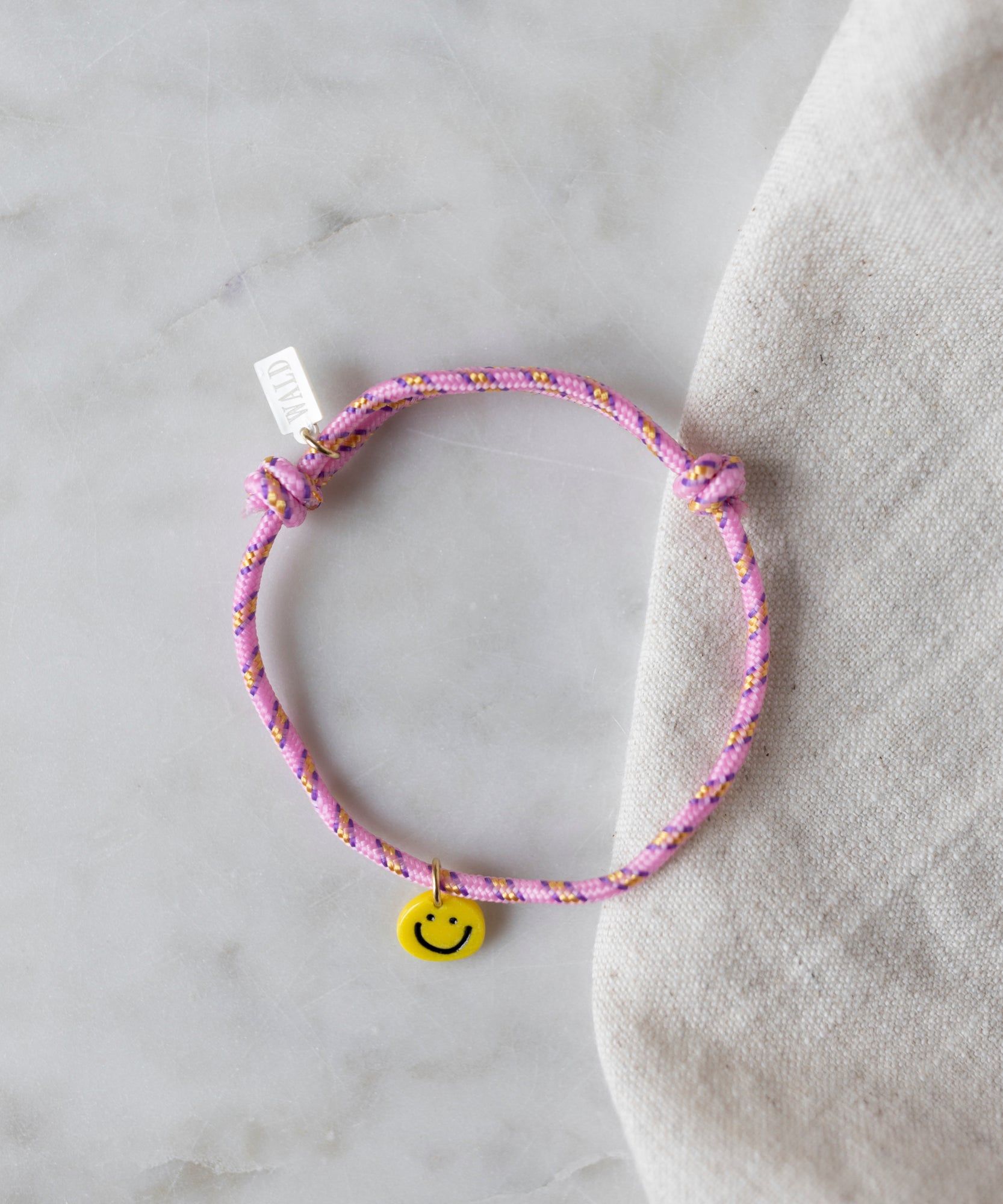 The Smilie Dude Pink Friendship Bracelet by WALD Berlin, featuring a yellow smiley face charm, is displayed on a marble surface.