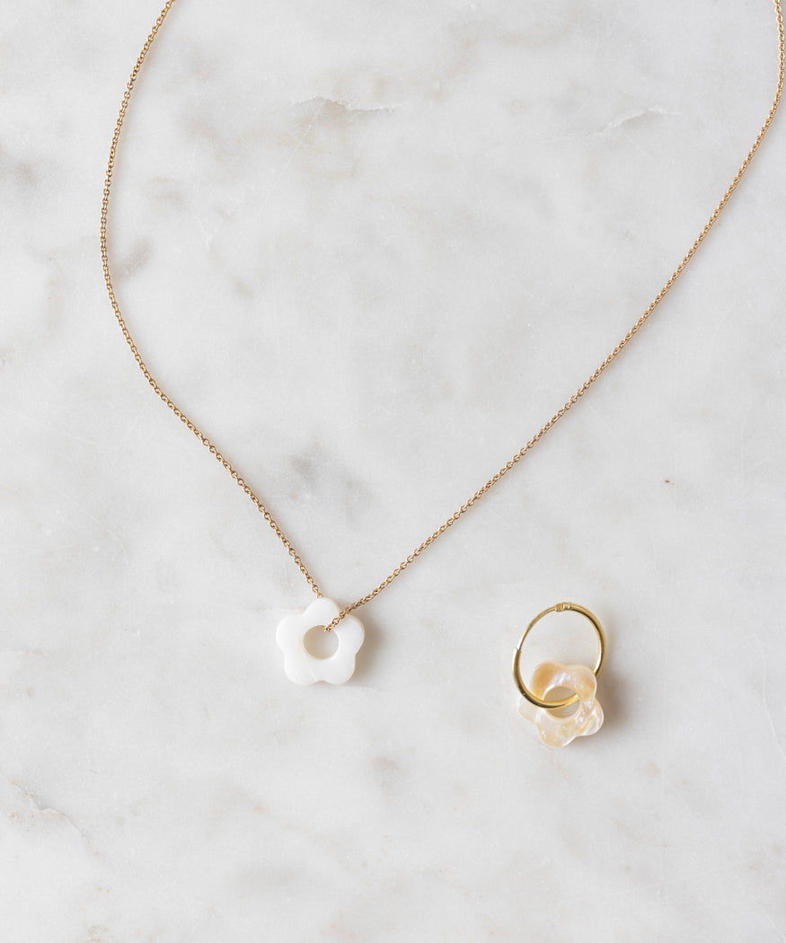 The WALD World Mother of Pearl Flower Charm Set, featuring a gold chain necklace adorned with a white flower-shaped pendant crafted from real mother of pearl flowers and a gold ring enhanced with an iridescent loop detail, is elegantly displayed on a marble surface.