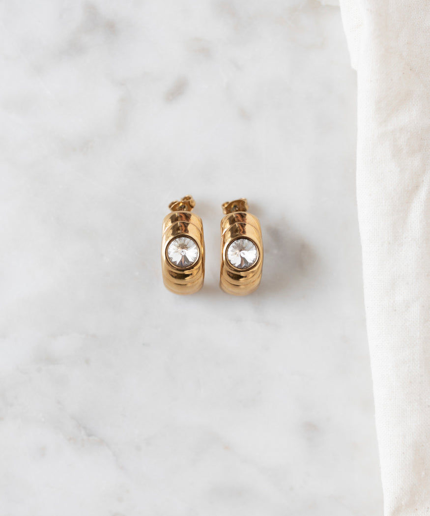 The Isla Bonita Ready or Not earrings by WALD Berlin, featuring a single clear Swarovski stone on each gold hoop, rest elegantly on a marble surface beside a white fabric, capturing an 80s vibe.