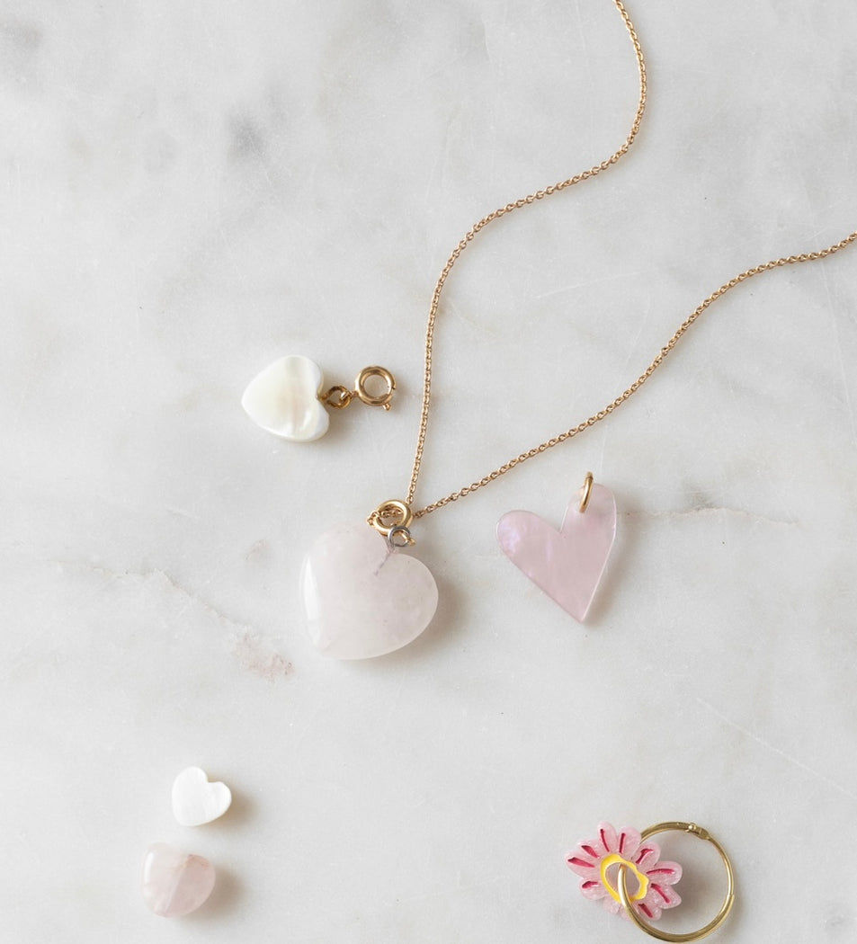 A gold necklace with a heart-shaped pendant, two separate heart pendants, a small flower ring, and a pair of tiny WALD World mother of pearl heart charm earrings are laid out on a white marble surface.