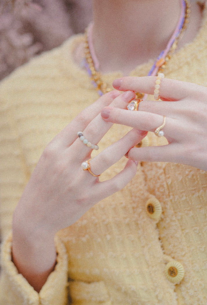 A person wears a yellow textured jacket, layered beaded necklaces, and several WALD Berlin NYC If I Rule The World Rings—colorful gemstone pieces hand-assembled from recycled gold—on their fingers.