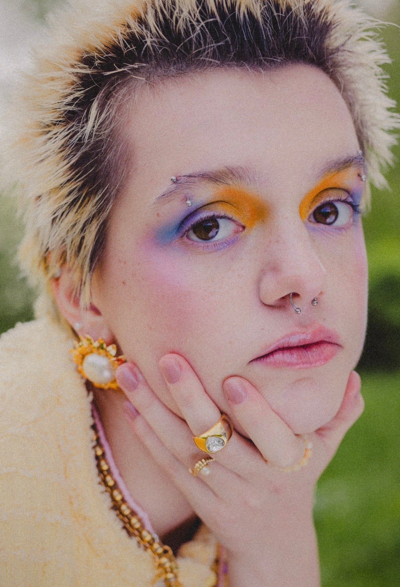 Person with short, spiky blonde hair and striking eye makeup poses outdoors, wearing WALD Berlin’s Pop Soleil Earrings in 18k gold plate along with gold jewelry.
