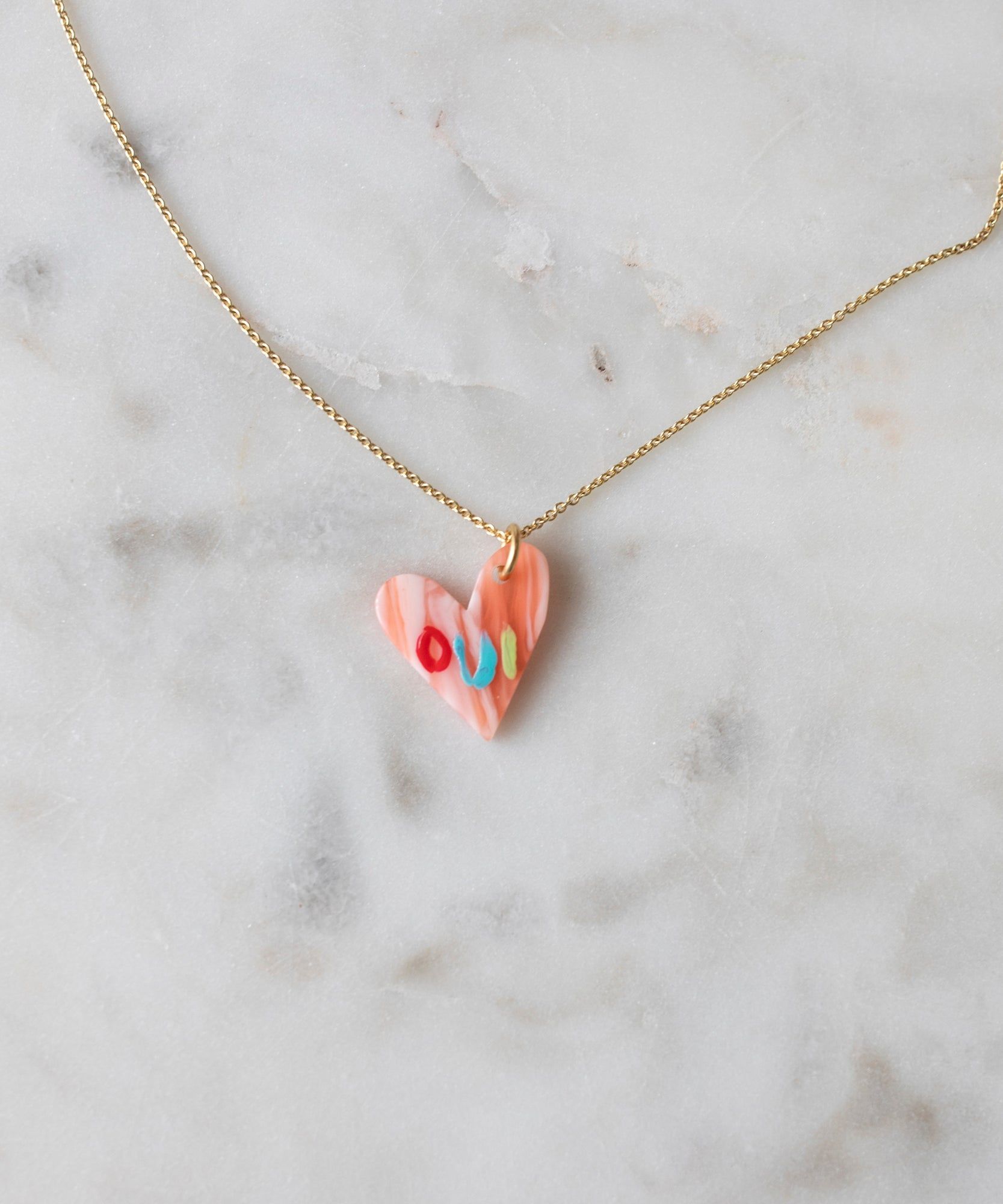 A colorful heart-shaped Pledges Of Tenderness LOVE Necklace made by WALD Berlin, on a gold chain, resting on a marble surface.