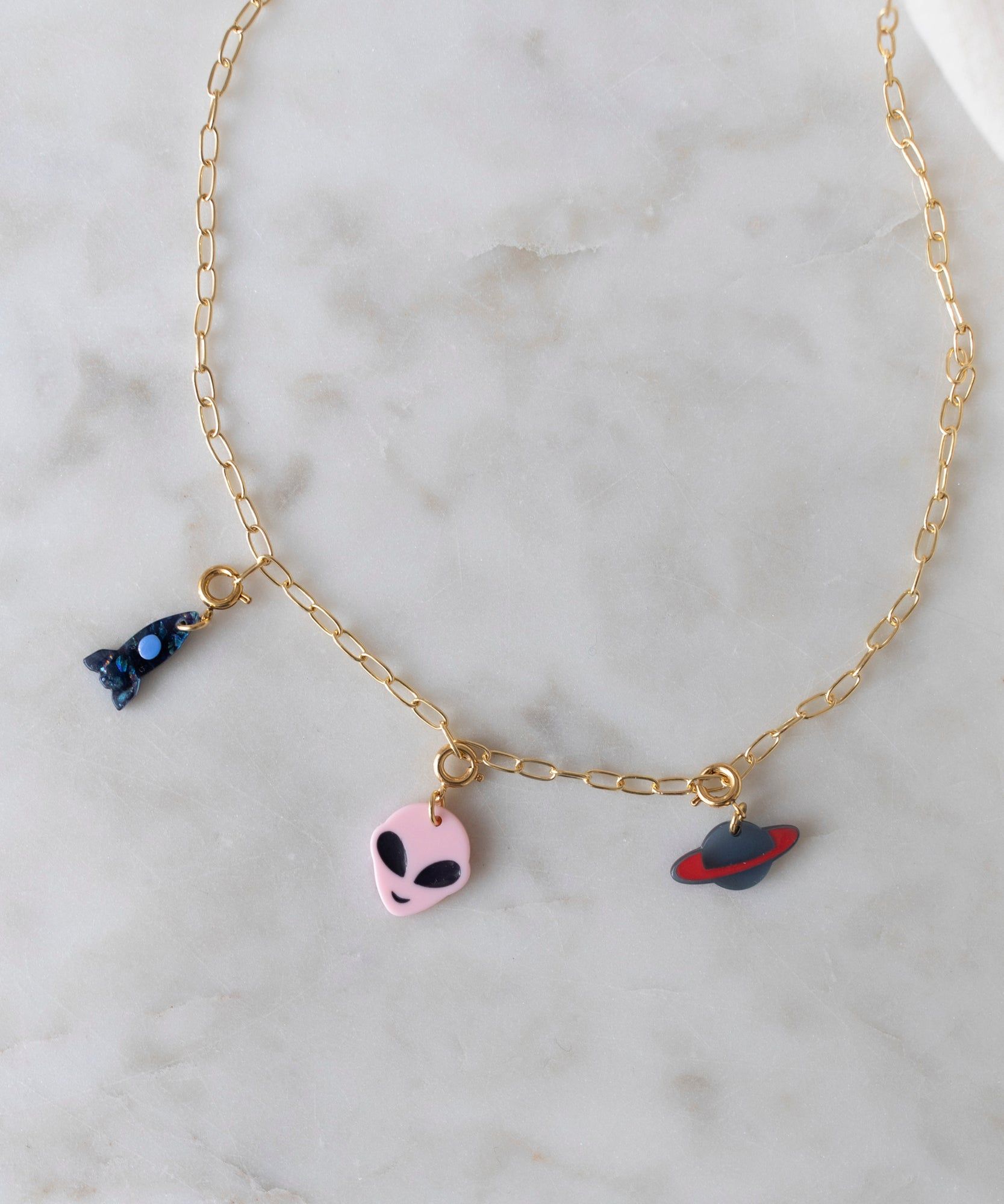 WALD Berlin Alien Dark Necklace with three colorful pendant charms: a pink alien face, a blue rocket, and a red planet, displayed on a marble surface. Made in Germany.