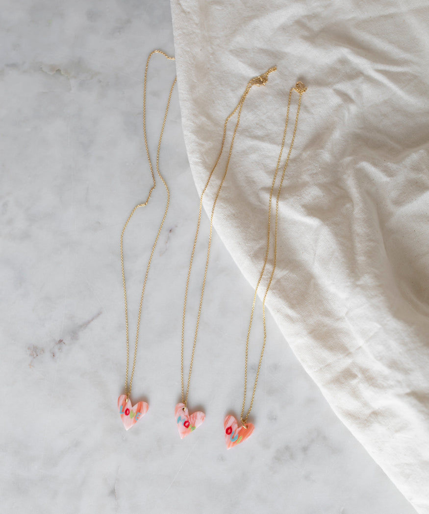 Three Pledges Of Tenderness MOM Necklaces with heart-shaped pendants on a marble surface, partially covered by a white cloth from WALD Berlin.