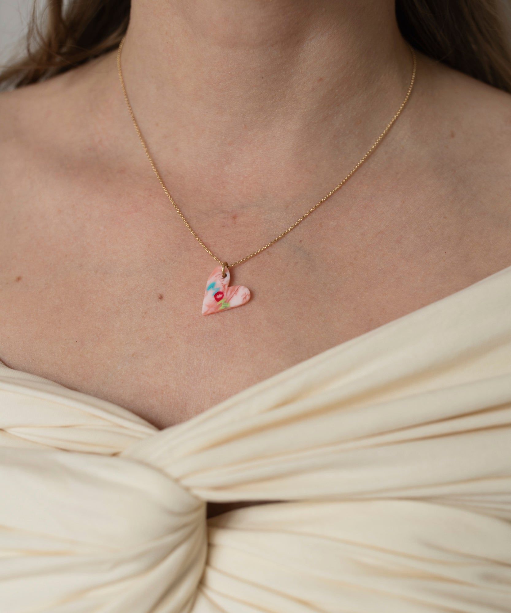 A close-up of a woman wearing a Pledges Of Tenderness LOVE Necklace by WALD Berlin, with a heart-shaped pendant, made in Germany, set against her cream dress.