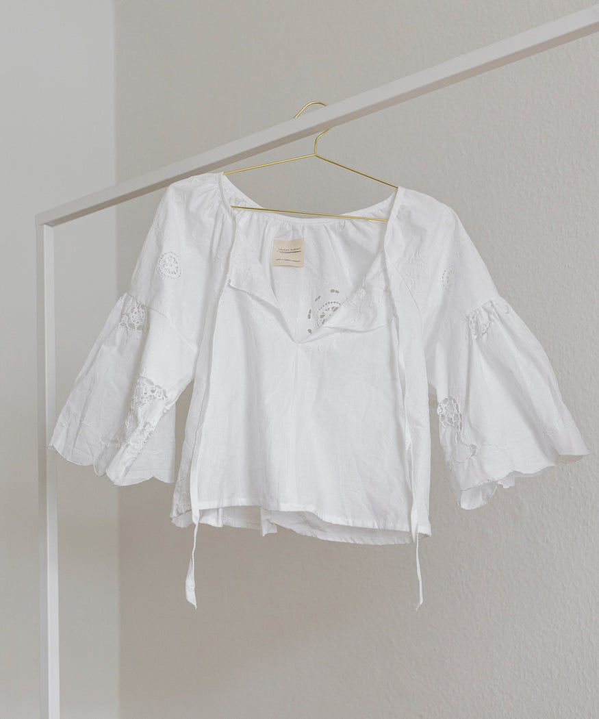 The WALD Berlin x Helena Harfst Necklace Blouse by WALD Berlin, a white handmade long-sleeve blouse with bell sleeves and a V-neckline, hangs on a gold hanger against a neutral background.
