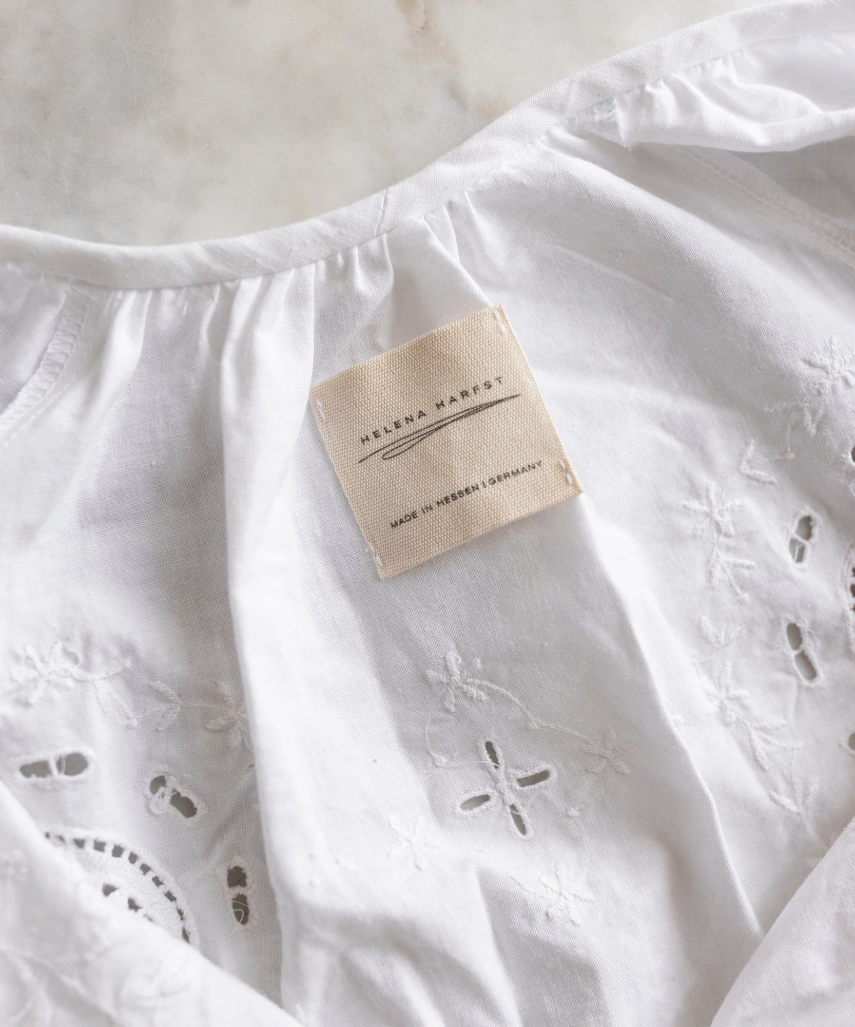 A close-up of the WALD Berlin x Helena Harfst Necklace Dress, a white garment with floral embroidery from a limited collection, featuring a beige label that reads 