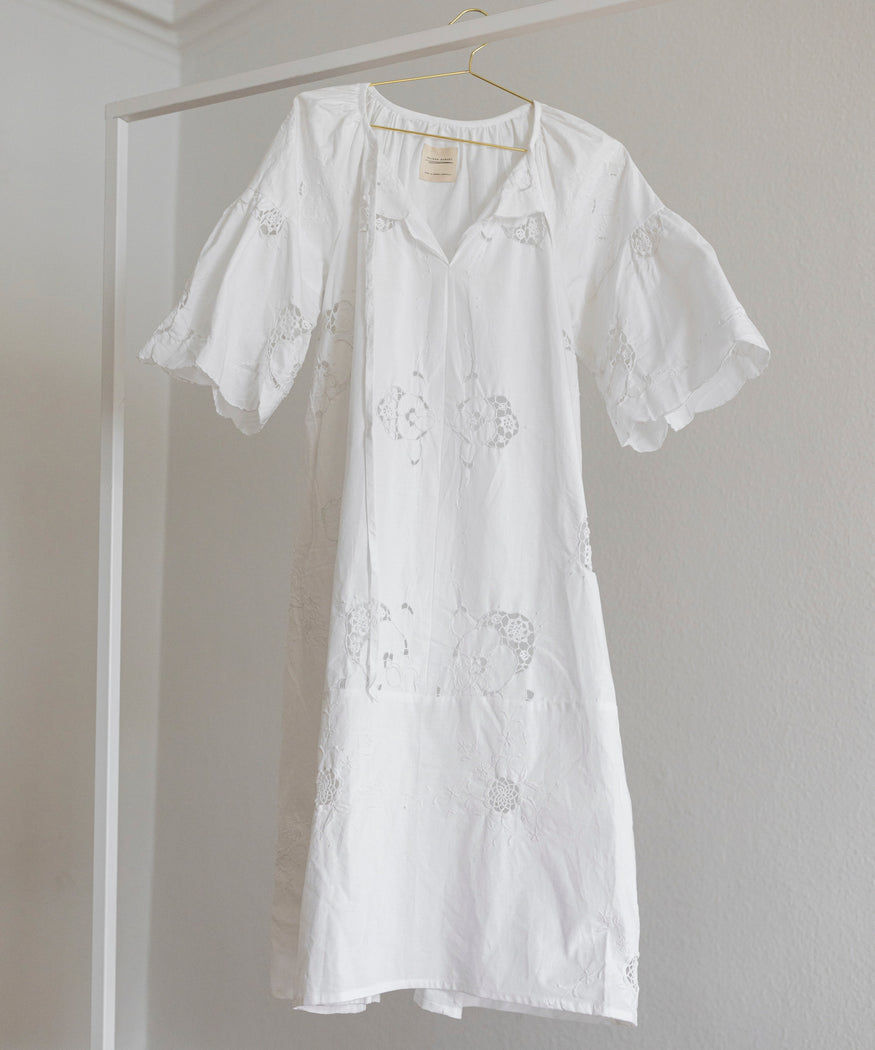 The WALD Berlin x Helena Harfst Necklace Dress, a white knee-length piece with short puffed sleeves and floral embroidery, from WALD Berlin’s limited collection, hangs on a hanger against a light-colored wall.