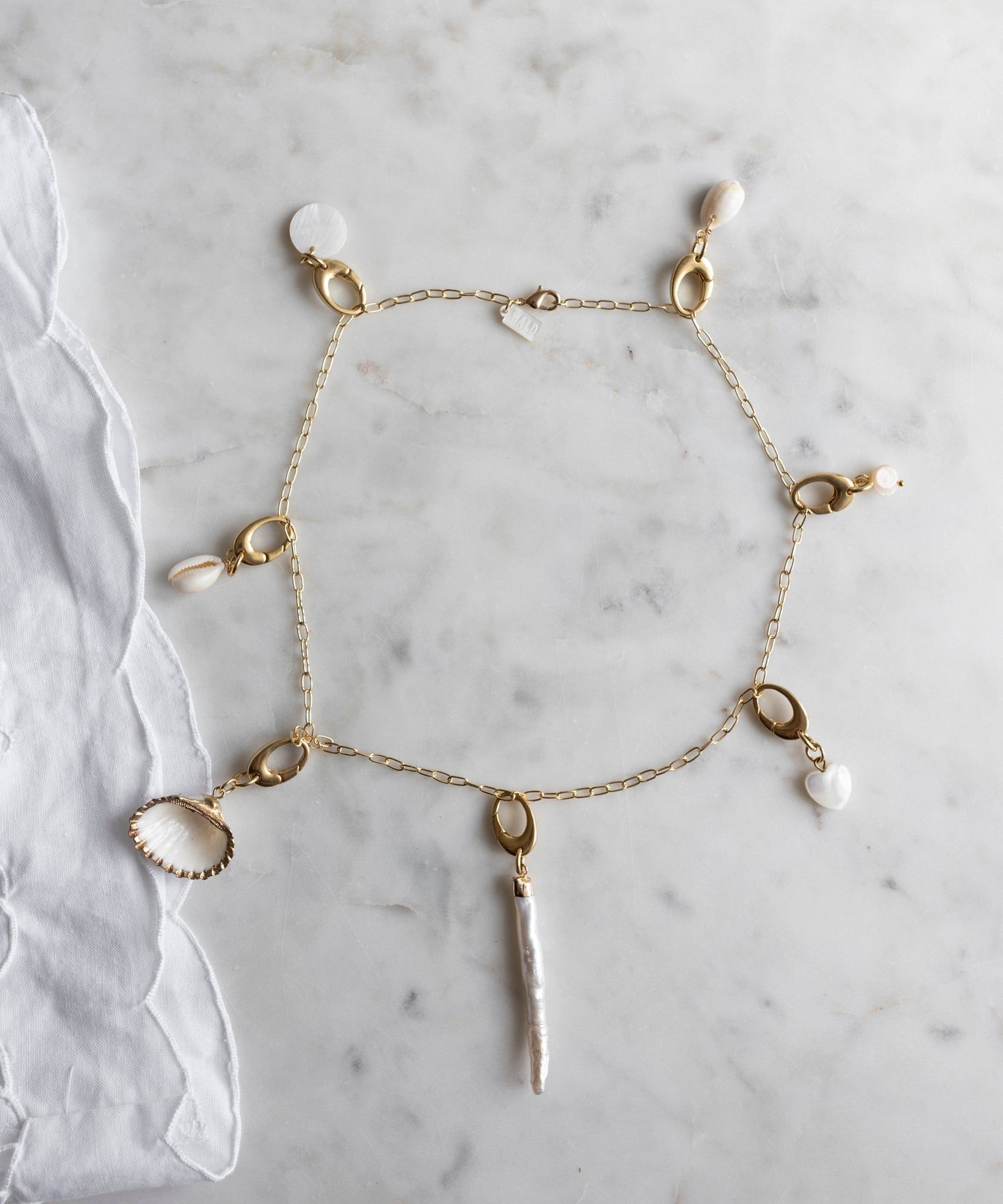 The WALD Berlin x Helena Harfst Necklace Dress by WALD Berlin is a limited collection gold charm necklace with seven dangling shells and pearls, shown on marble beside white fabric.