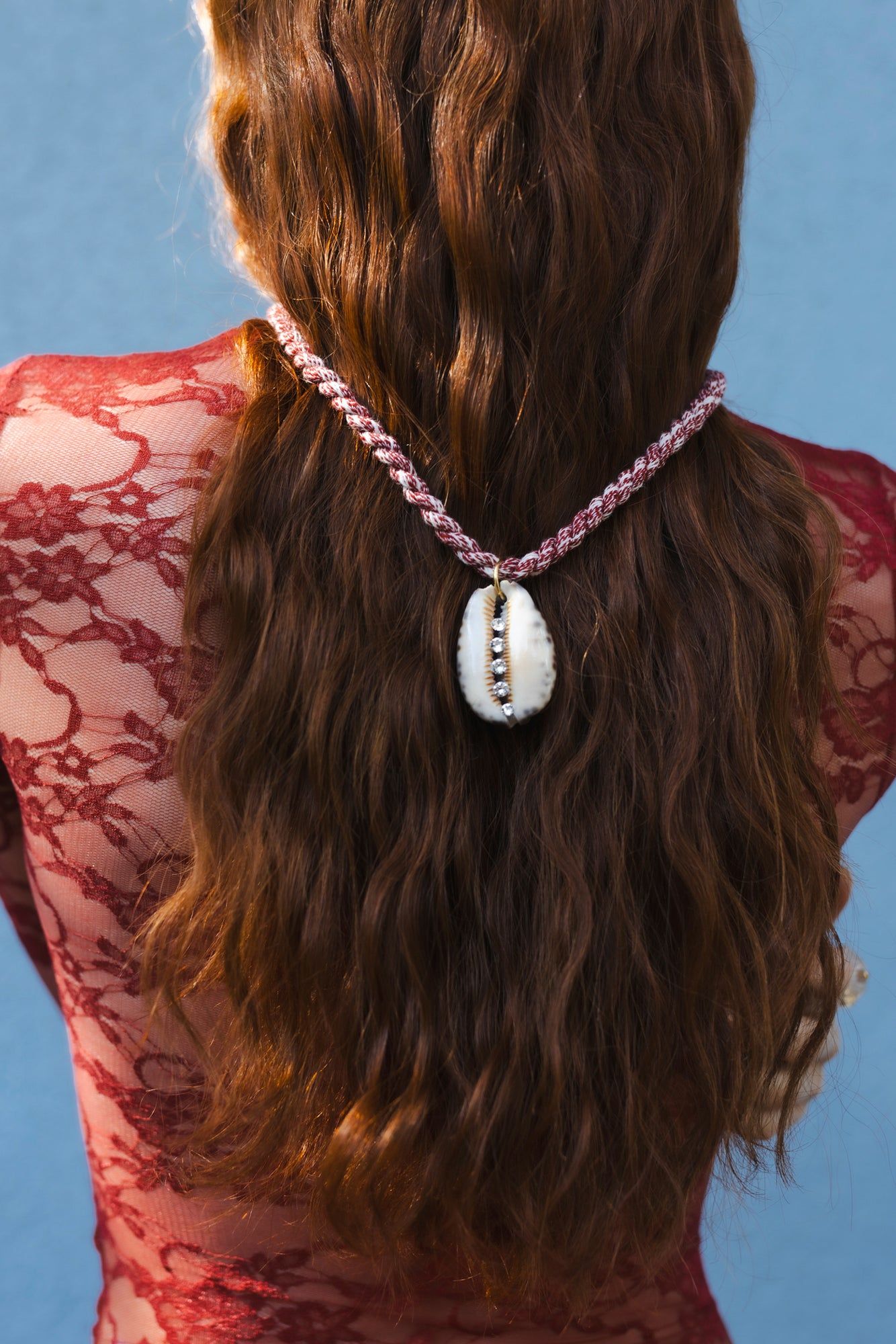 A woman with long, wavy brown hair and a red lace top faces away from the camera. She has the WALD Berlin La Piscina Bootylicious Necklace With Ribbon showcasing real shells and a Swarovski stone hanging down her back.